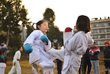 Two young children in martial arts uniforms spar outdoors, surrounded by other kids