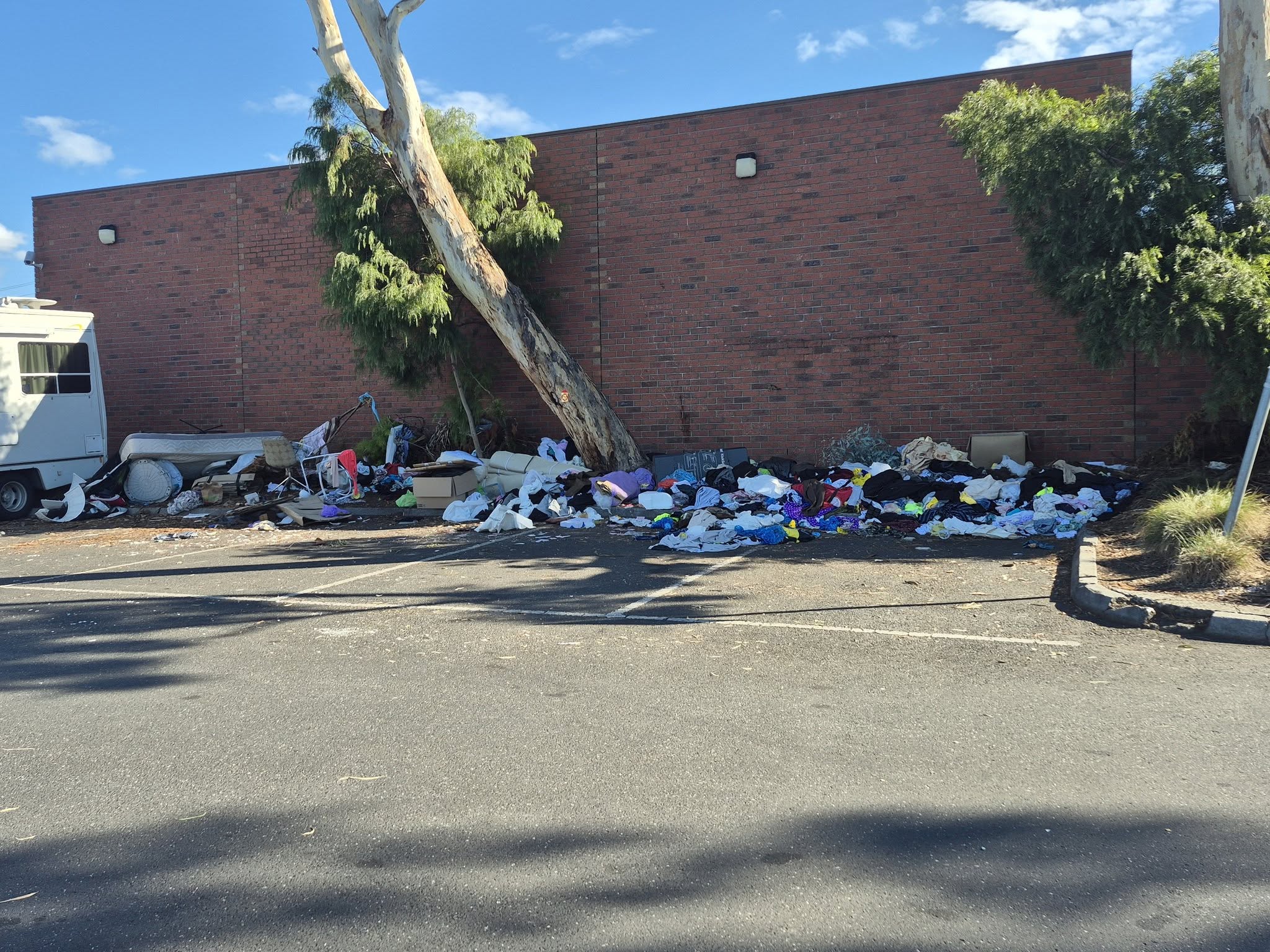 Rubbish and household items piled against a brick wall, in a carpark. 