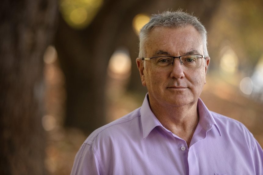 Portrait of a man with short grey hair and glasses in front of a blurry outdoor background