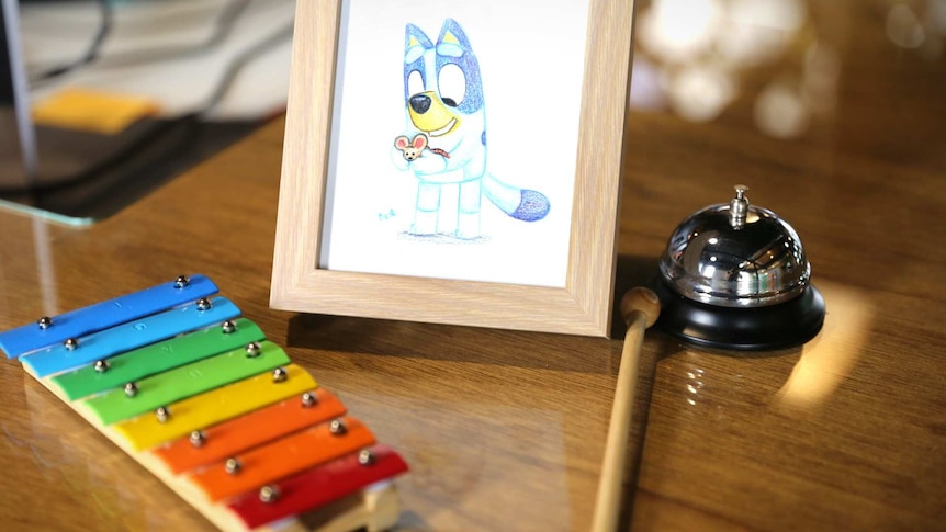 A desk with a coloured sketch of Bluey, a xylophone and a bell.