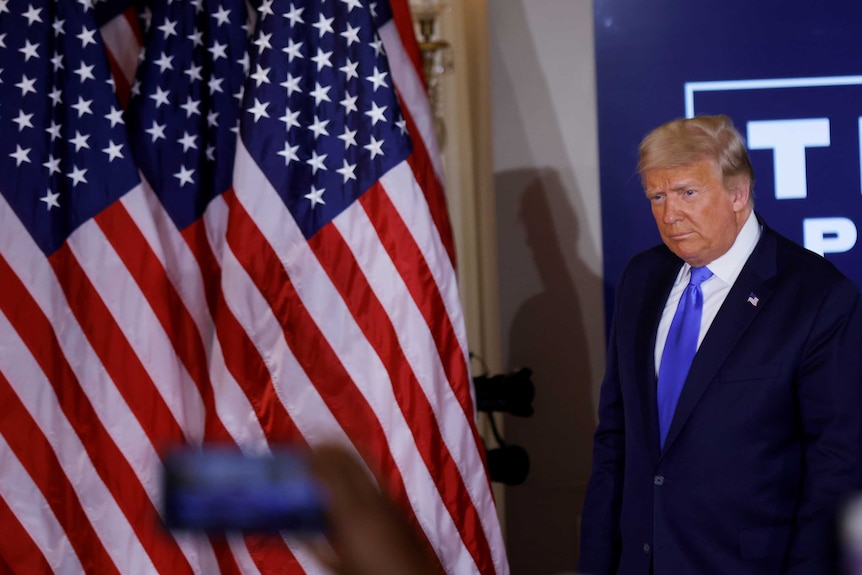 Donald Trump wearing a suit and blue tie walks in front of a stand of American flags.