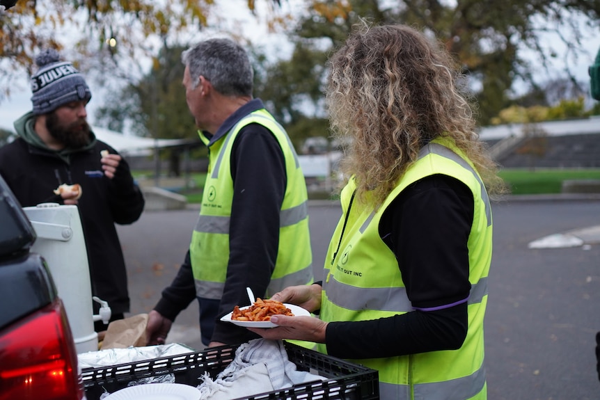 A woman in high-vis holding lasagna on a paper plate in a carpark.