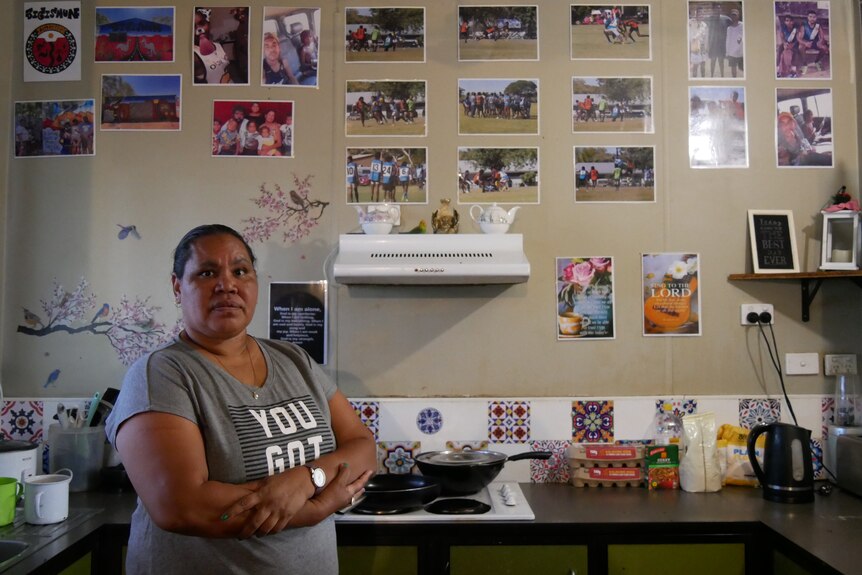 A woman standing with her arms cross in a kitchen with small photos covering the wall behind her.