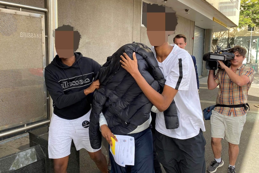 Two young men help someone walk while his face is covered by a black jacket to hide his identity from the media