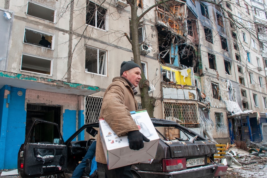 A man carries a box outside a damaged residential building, and near a damaged car.