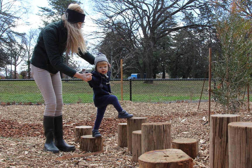 Child climbs onto a tree stump in a nature playground with his mum.