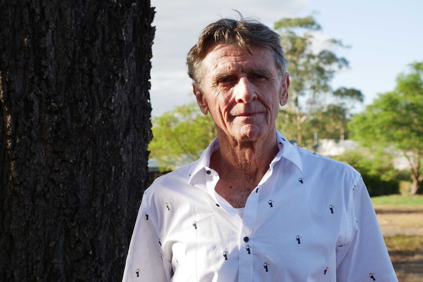 A fit-looking 70-year-old man wearing a white shirt stands next to a tree