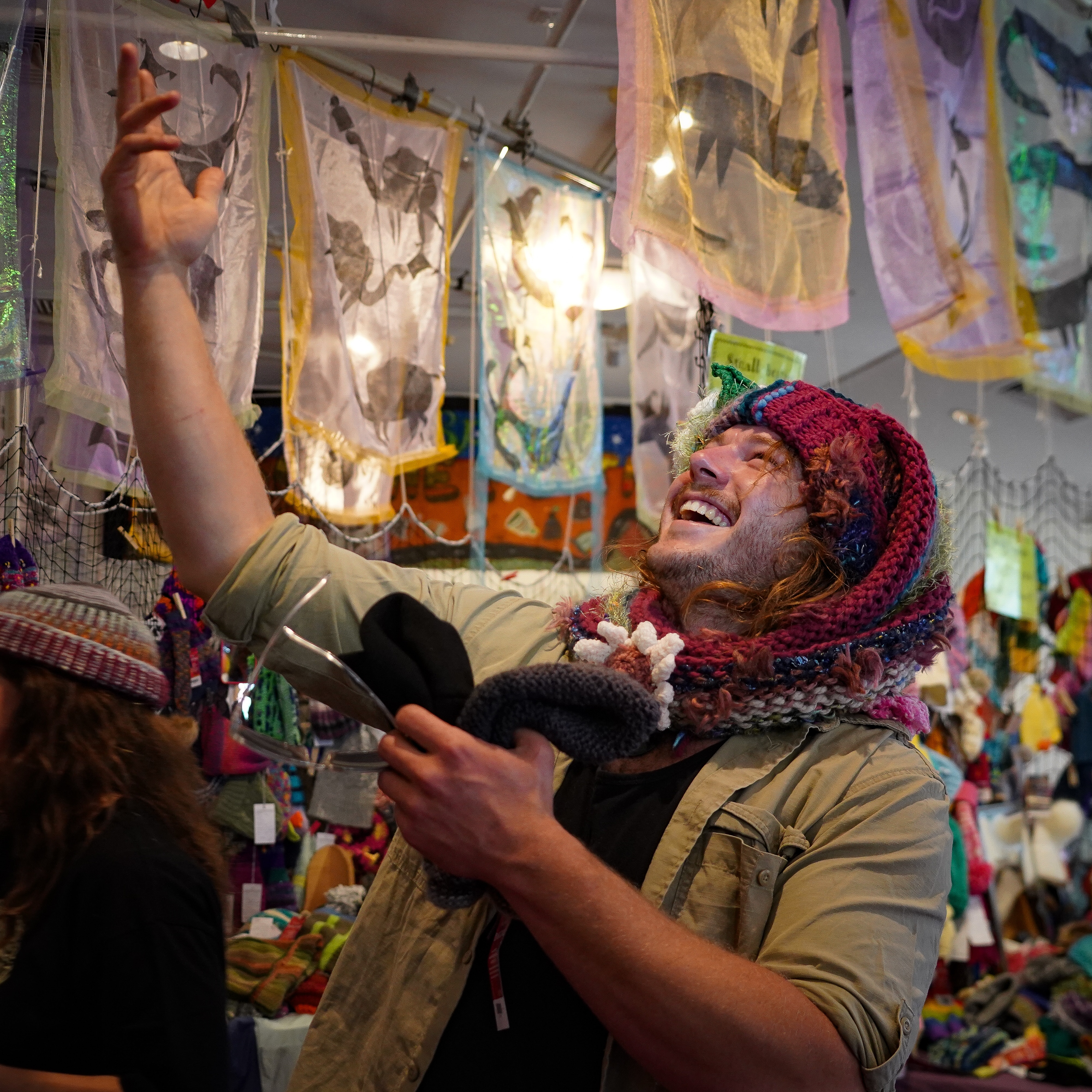 A man looking upwards and laughing, he is wearing a colourful handmade beanie with a knitted flower attached to it.