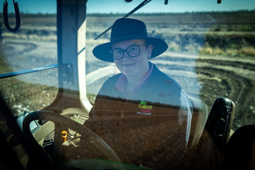 A women is pictured smiling through the windows of a tractor and she sits in the drivers seat behind the wheel. 