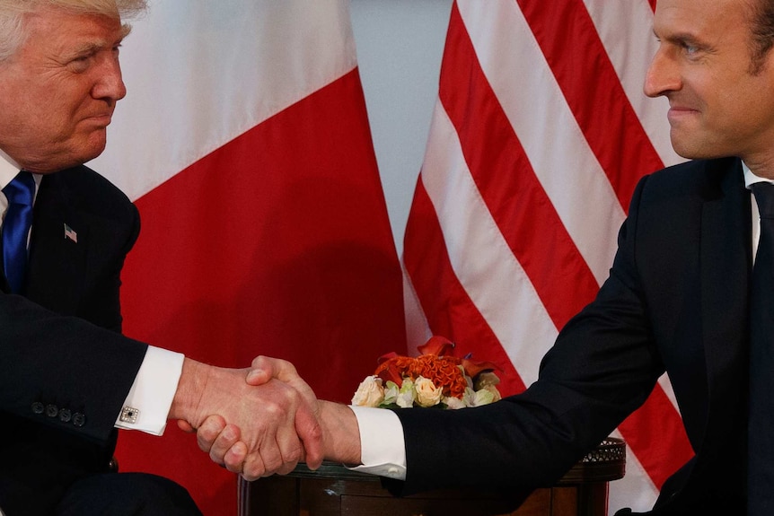 Trump (left) and Macon (right) grip each other's hands with purse-lipped smiles against a backdrop of French and US flags.
