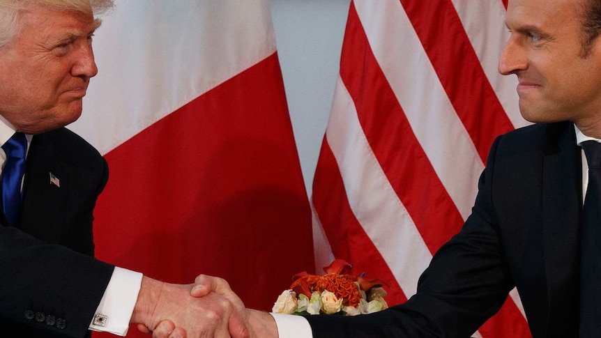 Trump (left) and Macon (right) grip each other's hands with purse-lipped smiles against a backdrop of French and US flags.