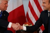 Trump (left) and Macon (right) grip each other's hands with purse-lipped smiles against a backdrop of French and US flags.