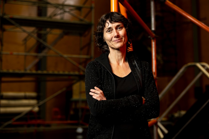 A middle-aged brunette woman stands with her arms folded in front of scaffolding. She is smiling slightly into the camera.