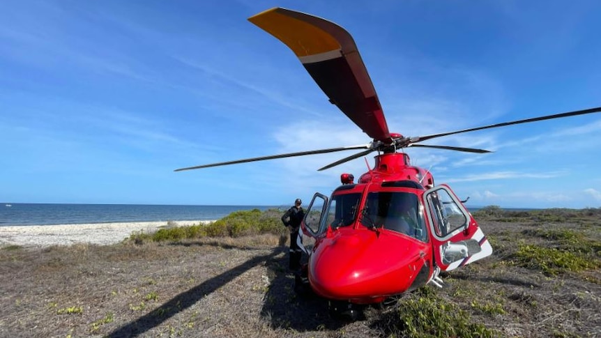 A man stands next to a red helicopter parked on an island, with the ocean on the left.