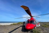 A man stands next to a red helicopter parked on an island, with the ocean on the left.
