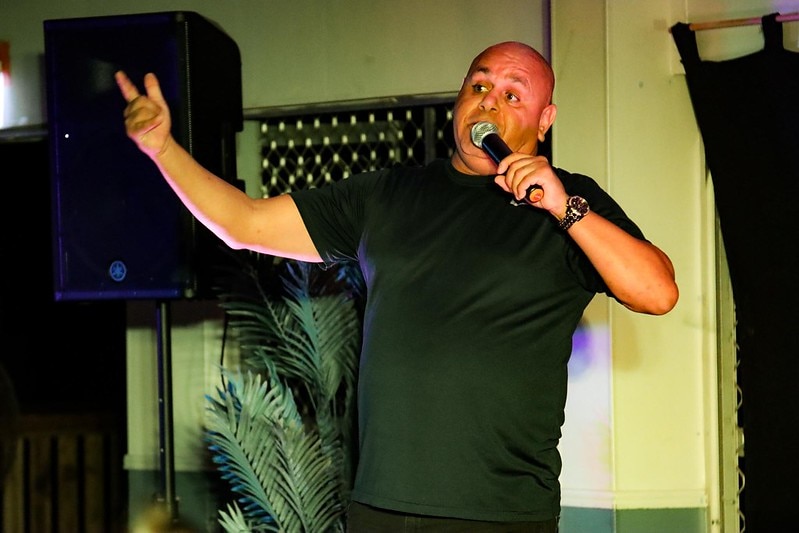 Bald Aboriginal male comedian performing stand-up, his right arm out-stretched with one finger pointing towards the crowd 
