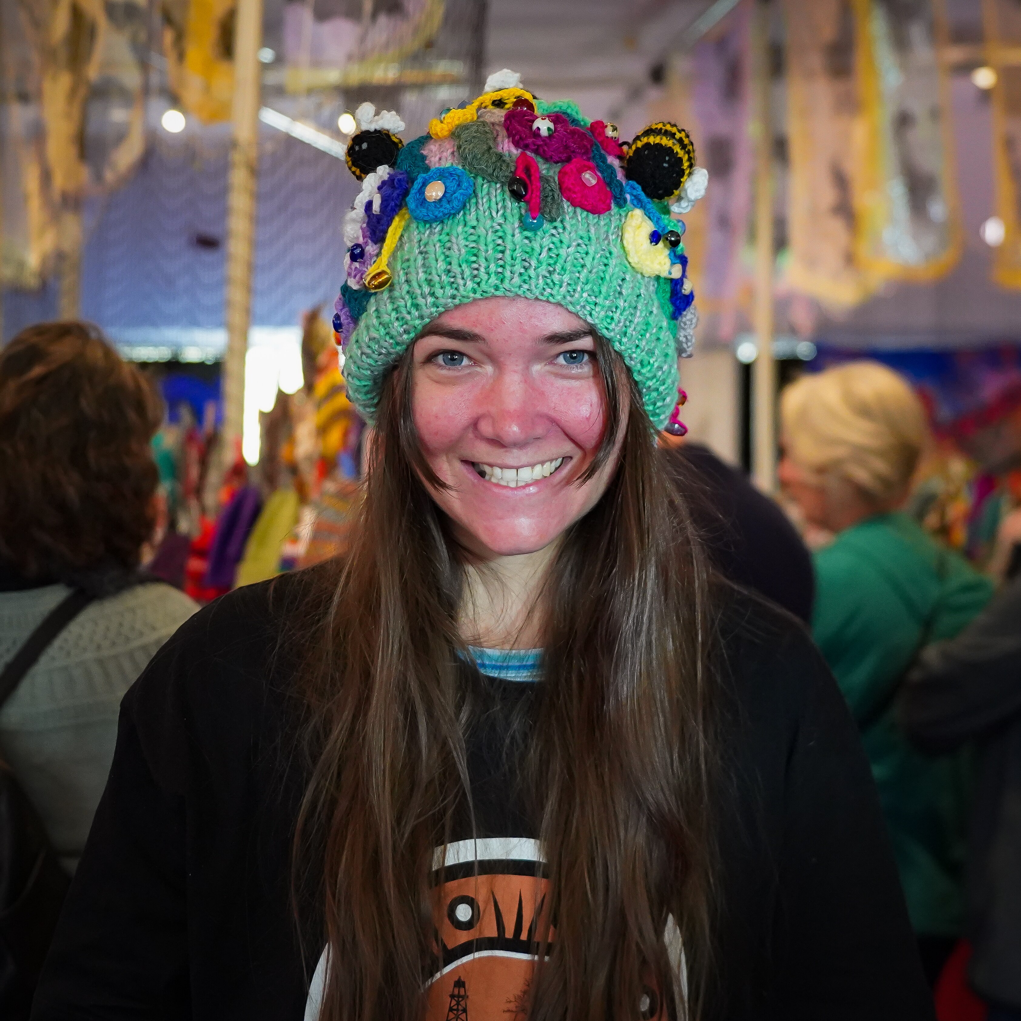 A woman smiles at the camera while wearing a green beanie, which has knitted flowers and bees attached to the top.