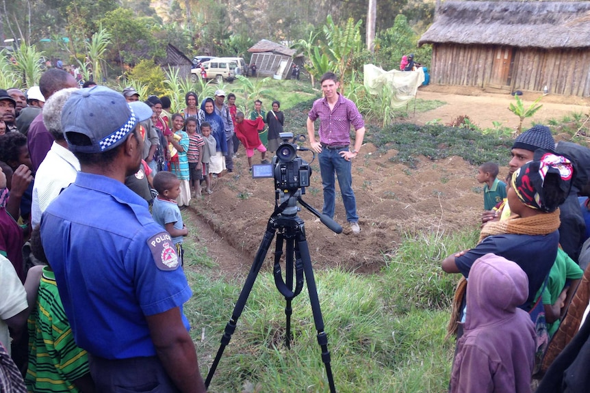 Tlozek standing in front of video camera surrounded by crowd in front of thatched hut and jungle.