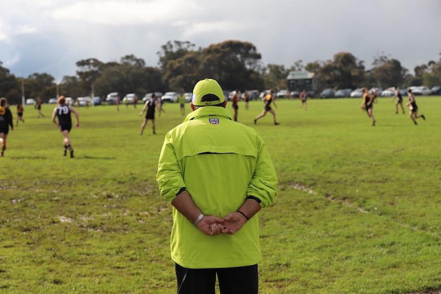 An umpire in a fluro outfit stands between goalposts looking out onto a football field. 