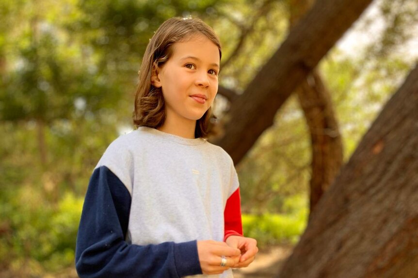 A young girl smiles as she stands by a tree.