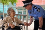 An elderly lady sits in a wheel chair and a police officer wearing a police hat holds her hand, they are both smiling.