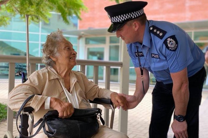 An elderly lady sits in a wheel chair and a police officer wearing a police hat holds her hand, they are both smiling.