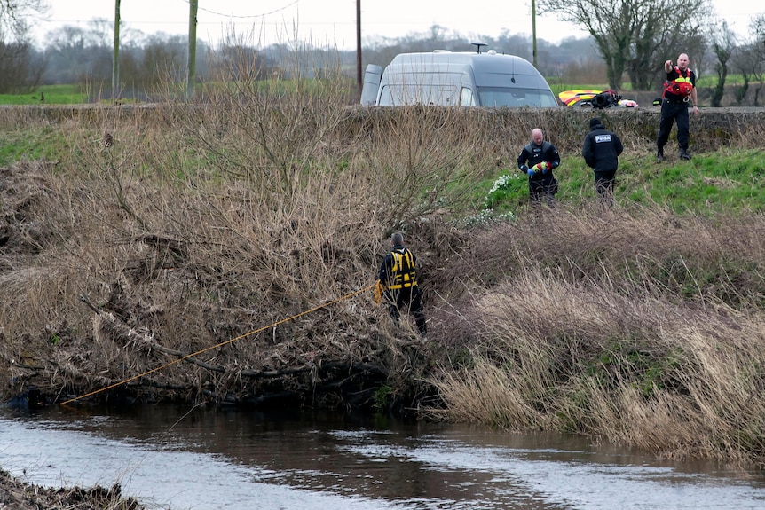 Police stand on a steep river bank covered in reeds