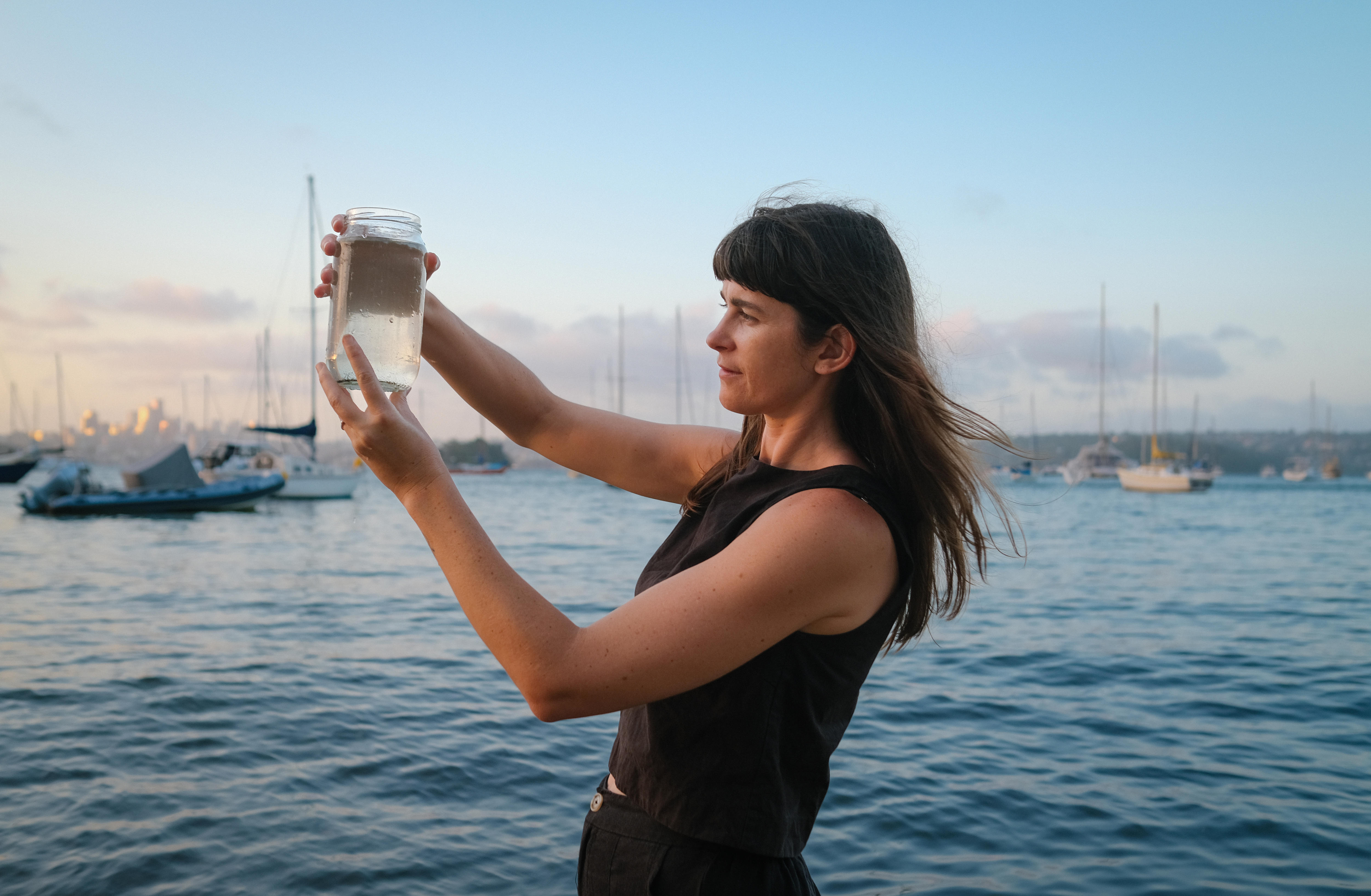 Sammy Hakwer collects harbour water in jar, holding it up high in front of her face at dusk 