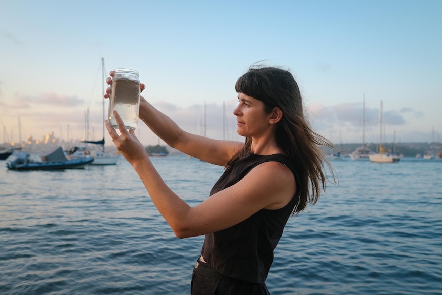 Sammy Hawker collects harbour water in jar, holding it up high in front of her face at dusk.