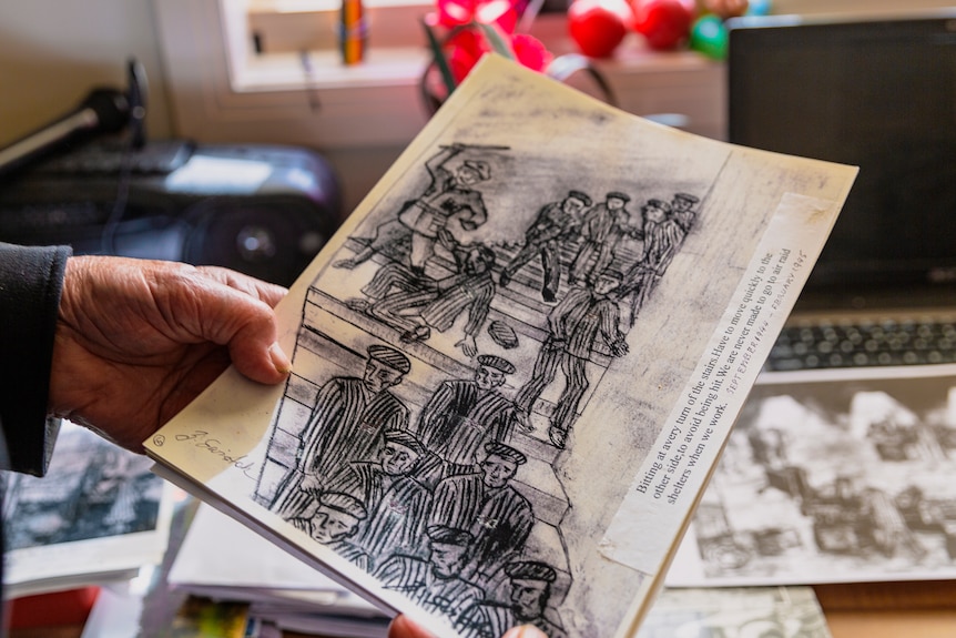 A hand holds out a black-and-white drawing of prisoners in a concentration camp.