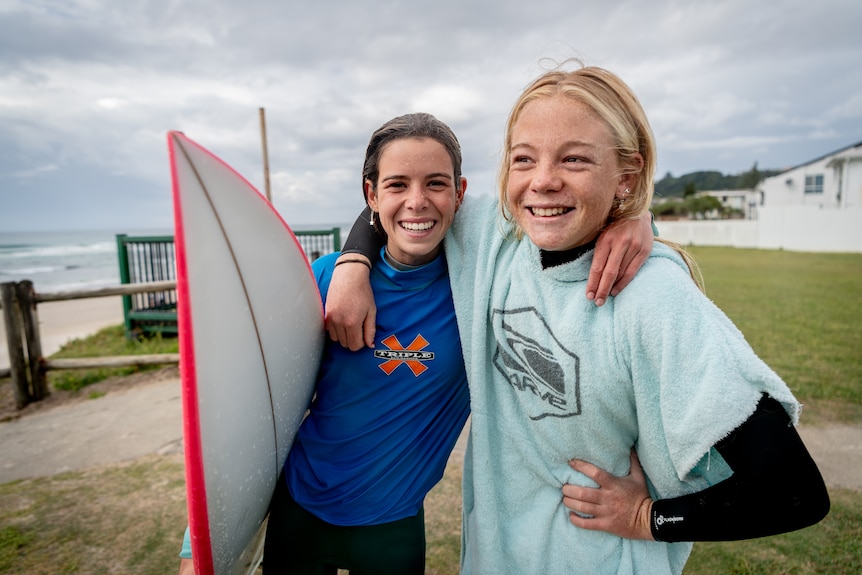 Two young girls smile arm-in-arm, one has a surfboard under her arm