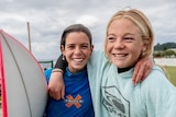 Two young girls smile arm-in-arm, one has a surfboard under her arm