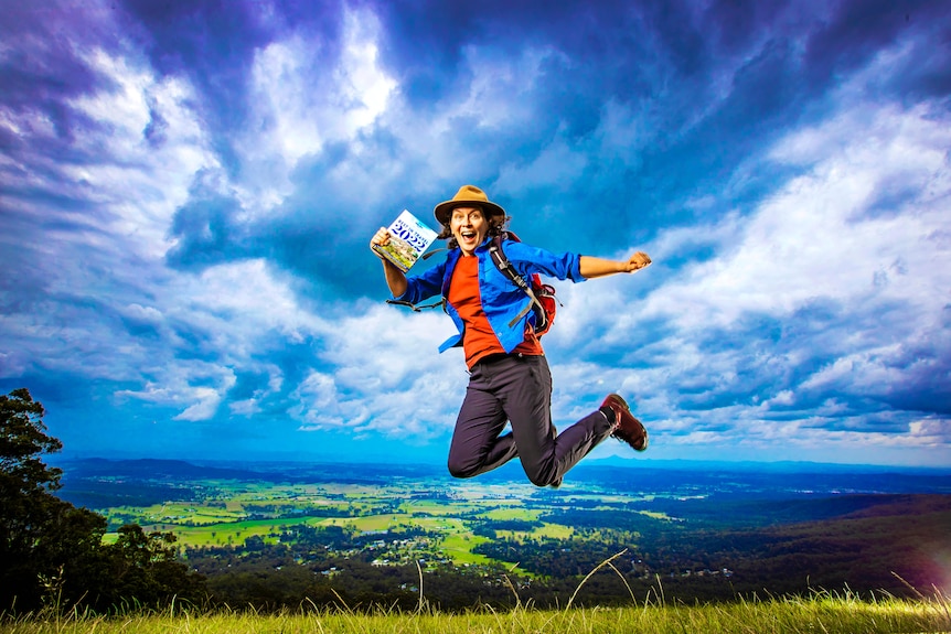 Woman holding a book and jumping into air on a hill top. 