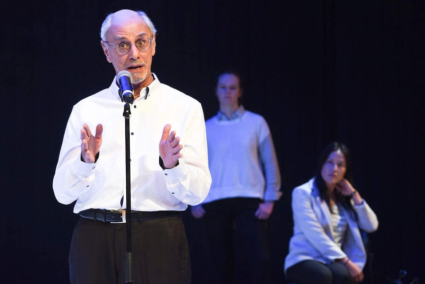 Three actors on a stage, two women in the background and a man with glasses in the front speaking into a microphone