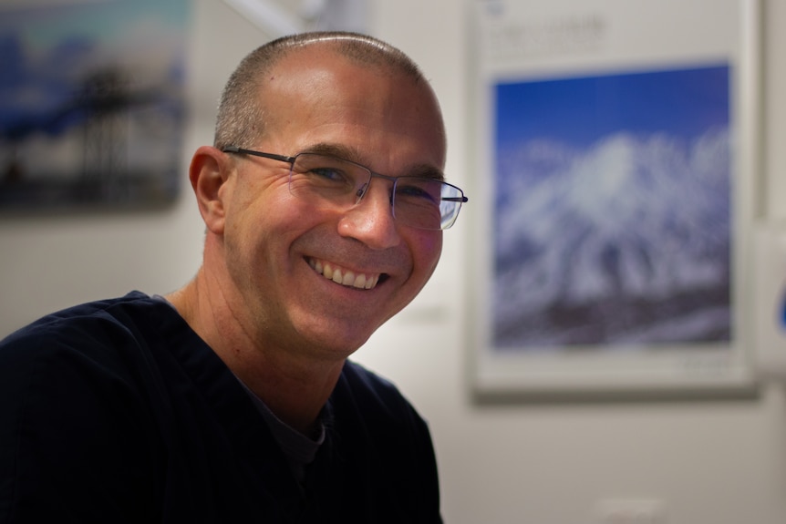 A headshot of a doctor smiling wearing blue scrubs in his office