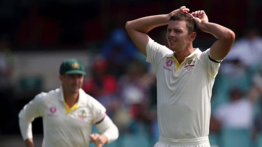 Australia bowler Josh Hazlewood puts his hands on his head with a pained expression. Shaun Marsh, blurred, runs in the field.