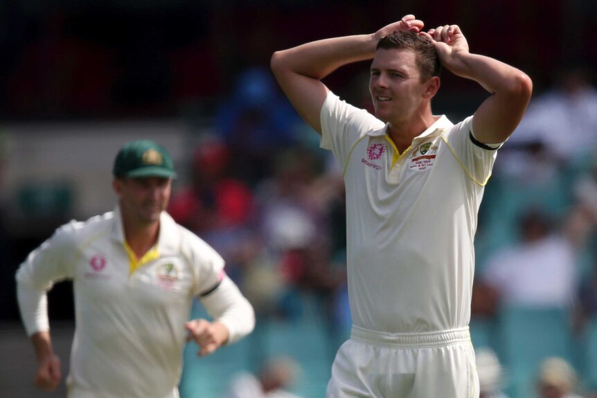 Australia bowler Josh Hazlewood puts his hands on his head with a pained expression. Shaun Marsh, blurred, runs in the field.