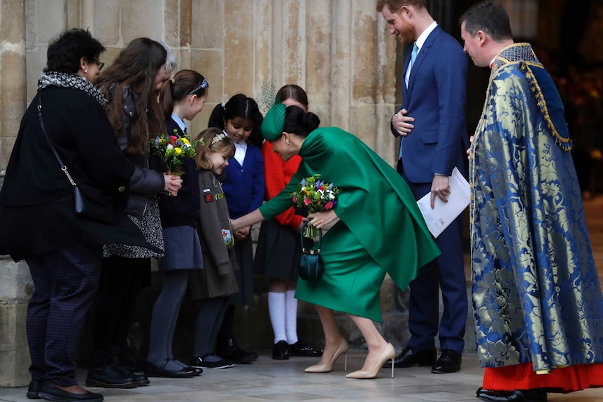 Meghan bends down to greet children as Prince Harry stands behind her.