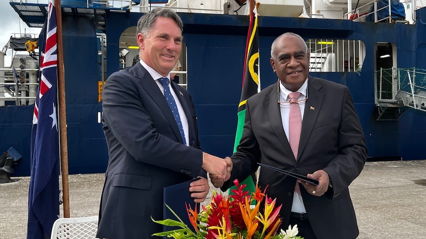 Richard Marles and Ishmael Kalsakau smile as they shake hands while standing in front of the flags of Vanuatu and Australia.