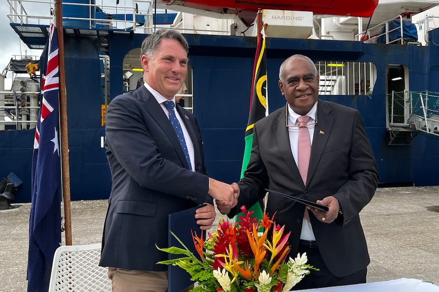 Richard Marles and Ishmael Kalsakau smile as they shake hands while standing in front of the flags of Vanuatu and Australia.