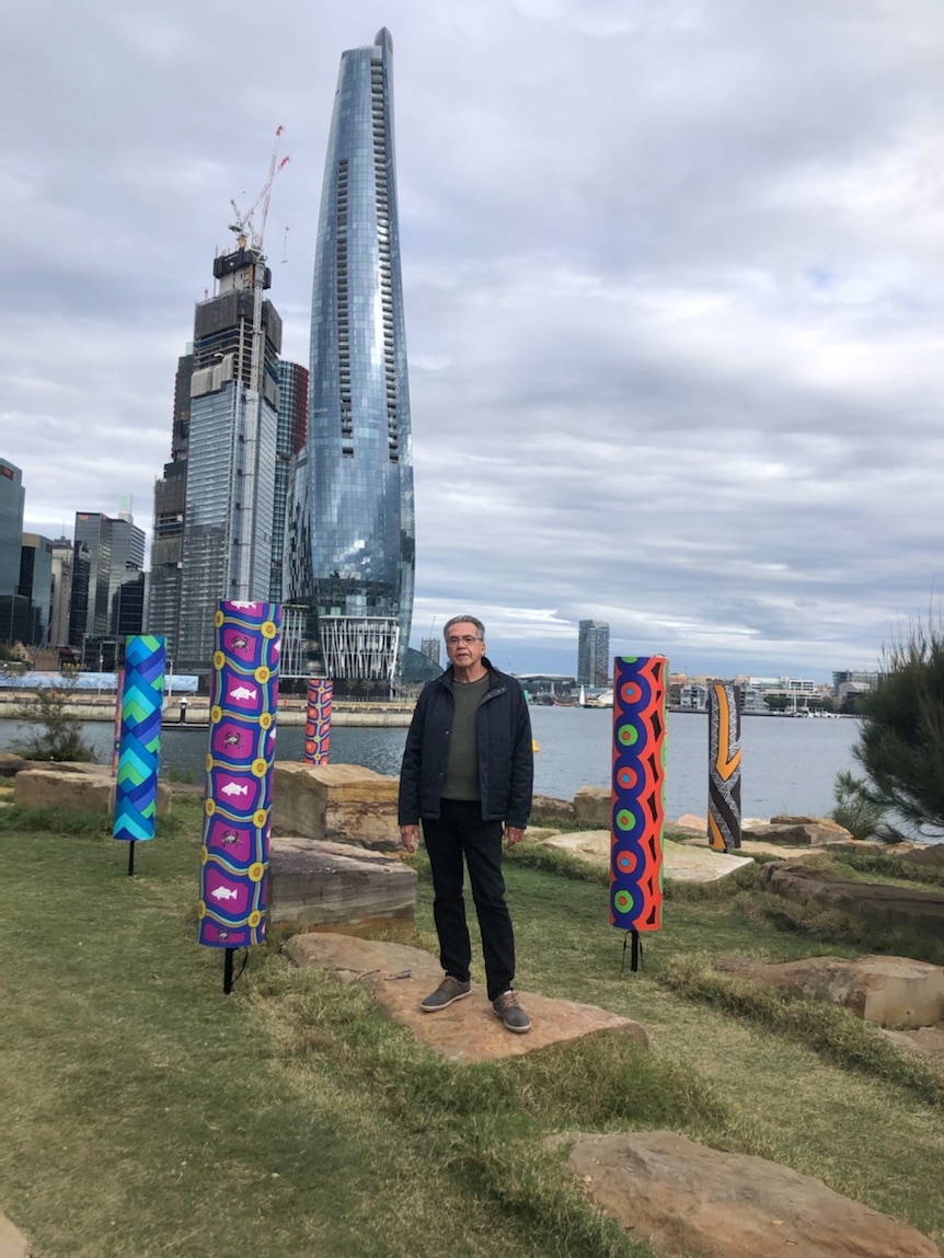 Warwick Keen stands wearing black alongside cylinders with his artwork on them and Sydney skyline in the background.
