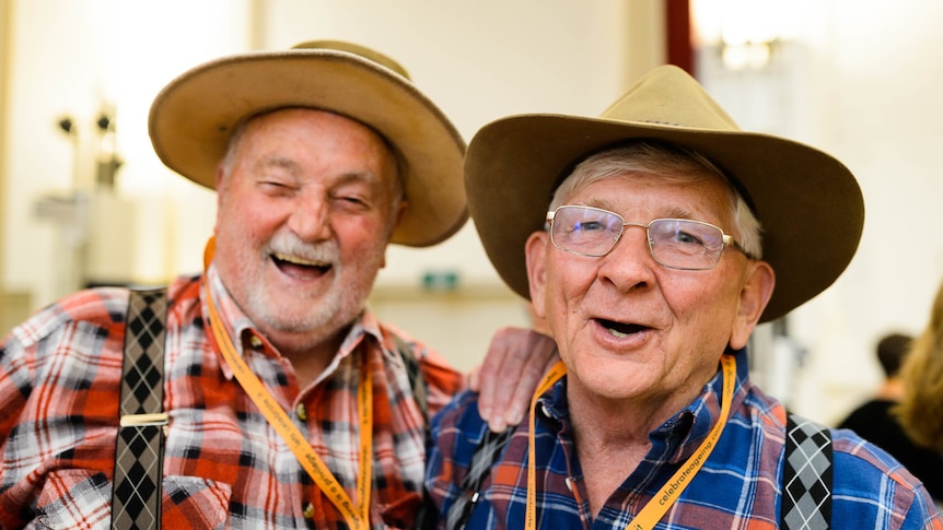 Two men in hats and flannelette shirts, one red and one blue, stand close together beaming widely.