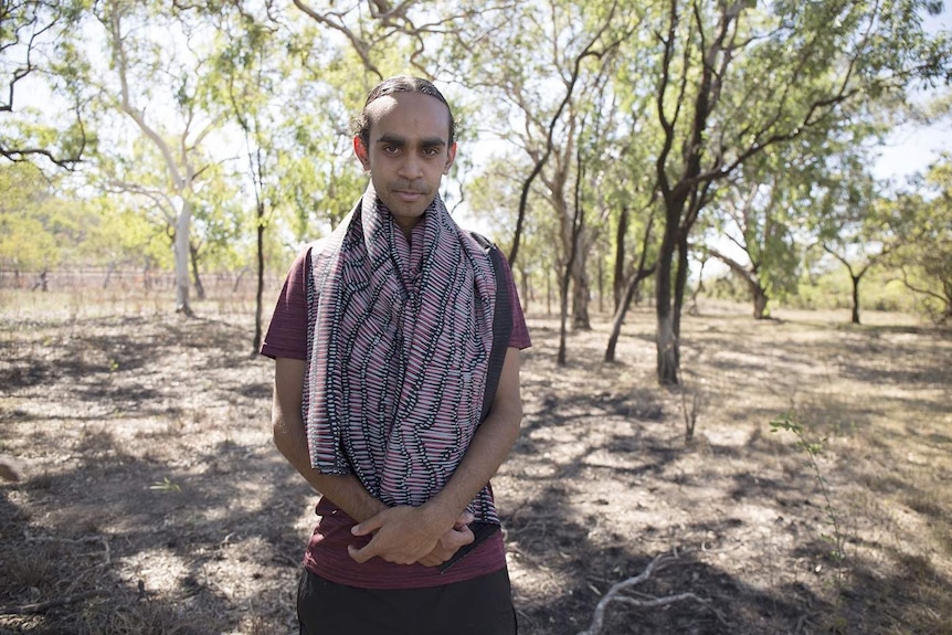 A man stands wearing a large piece of printed fabric over his shoulders in the NT bush.