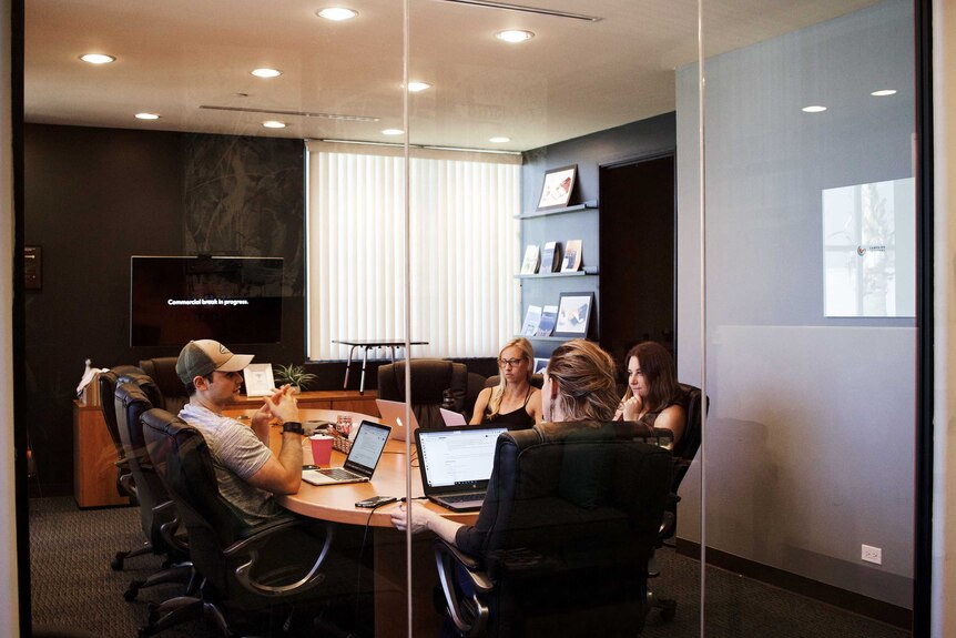 A group of young people sit around a table with their laptops in a conference room.