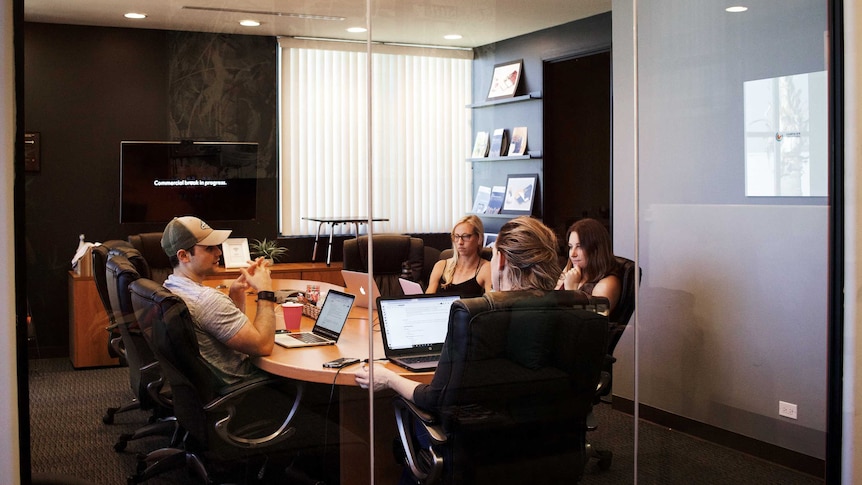 A group of young people sit around a table with their laptops in a conference room.