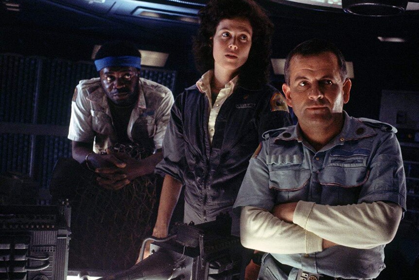 Yaphet Kotto, Sigourney Weaver and Ian Holm standing next to each other and looking off screen on the set of Alien