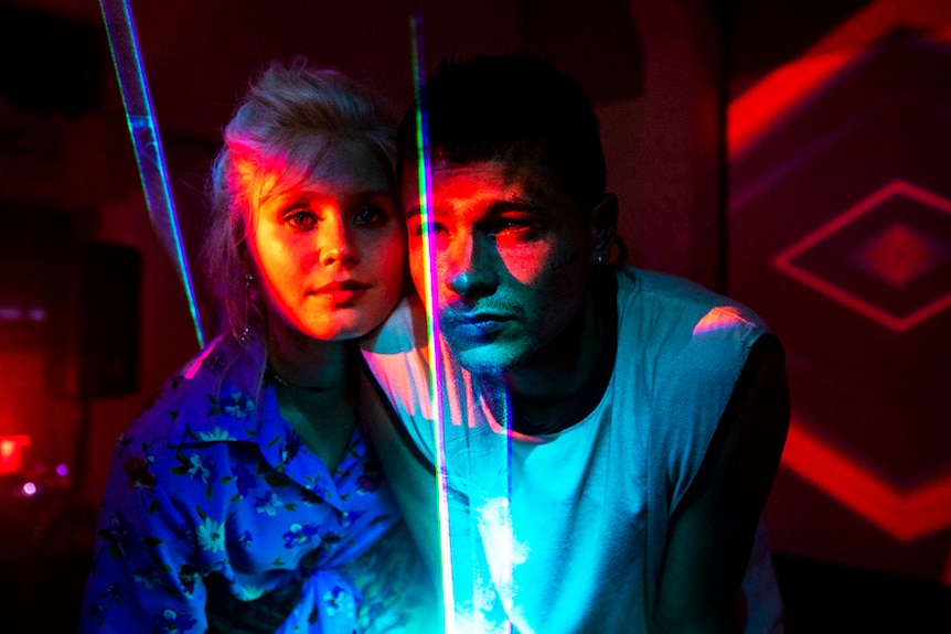 In a dark room near speaker a young girl and boy sit together, their faces and bodies illuminated by orange red and blue lasers.