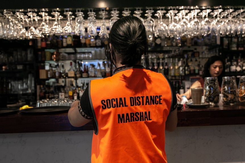 A woman stands with her back to the camera. She is wearing a vest with the words "covid safety marshal" on it.