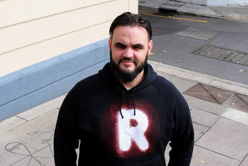 A man wearing a jumper with a Recognise logo looks at the camera from a city street.