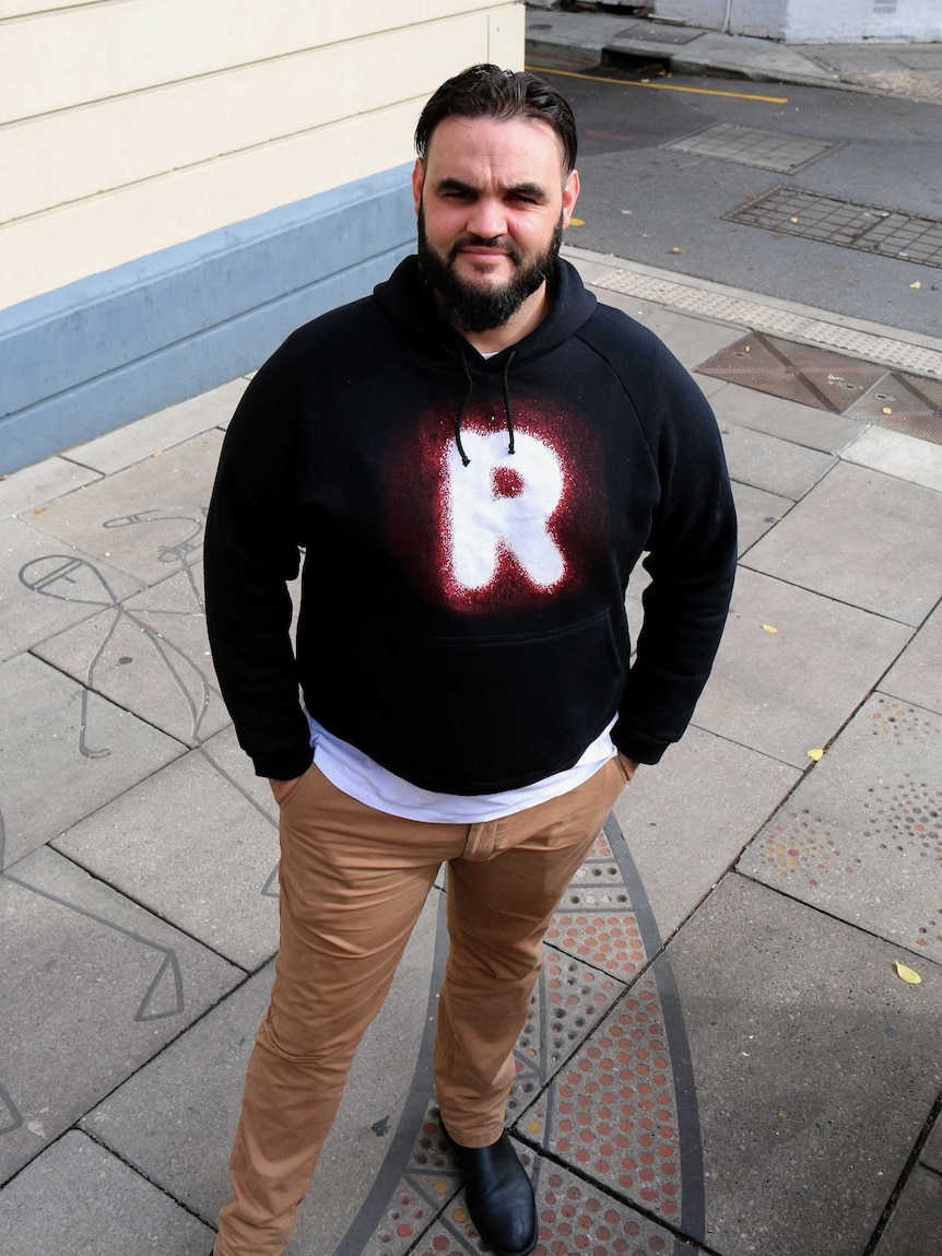 A man wearing a jumper with a Recognise logo looks at the camera from a city street.